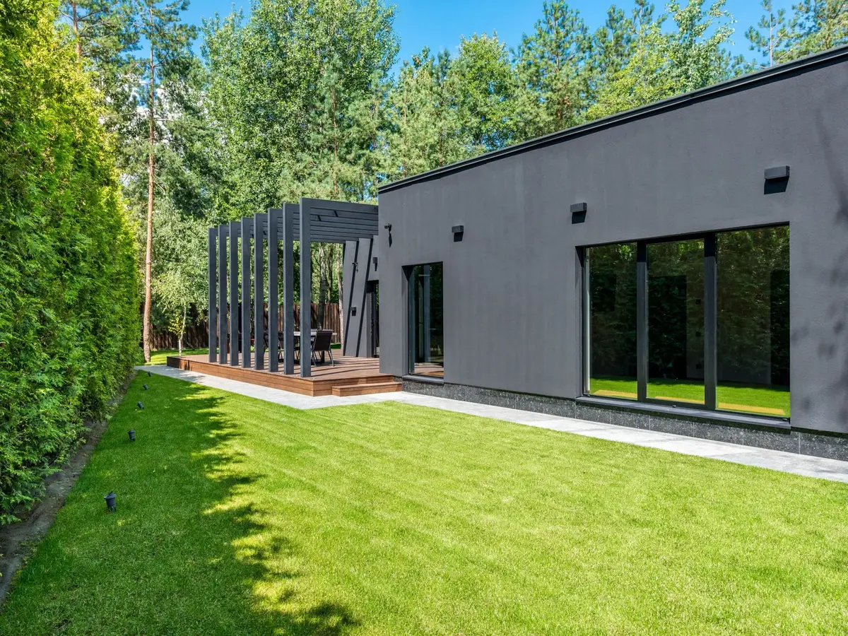 A green lawn between a tall hedge and a modern house, with pines behind