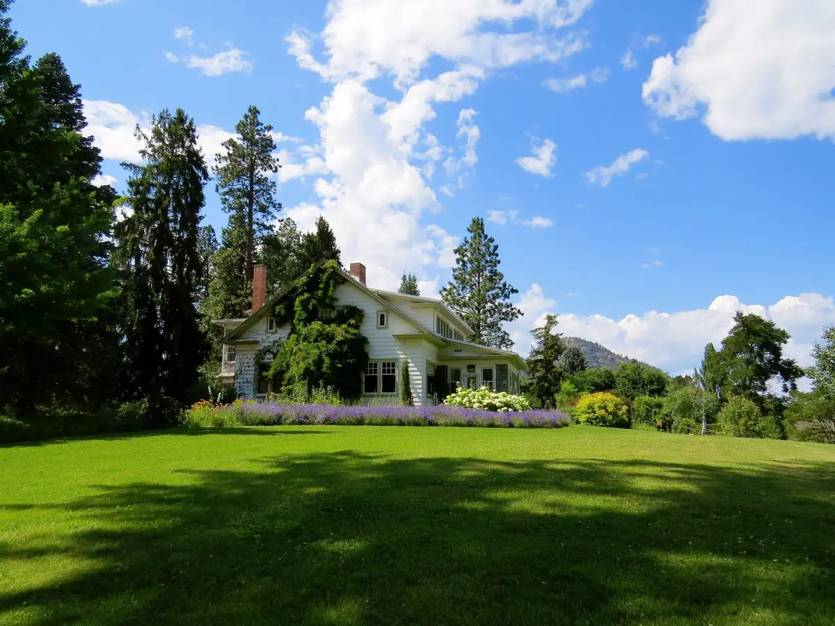 A wide green lawn in front of a white house, with tall trees and flower beds under a blue sky