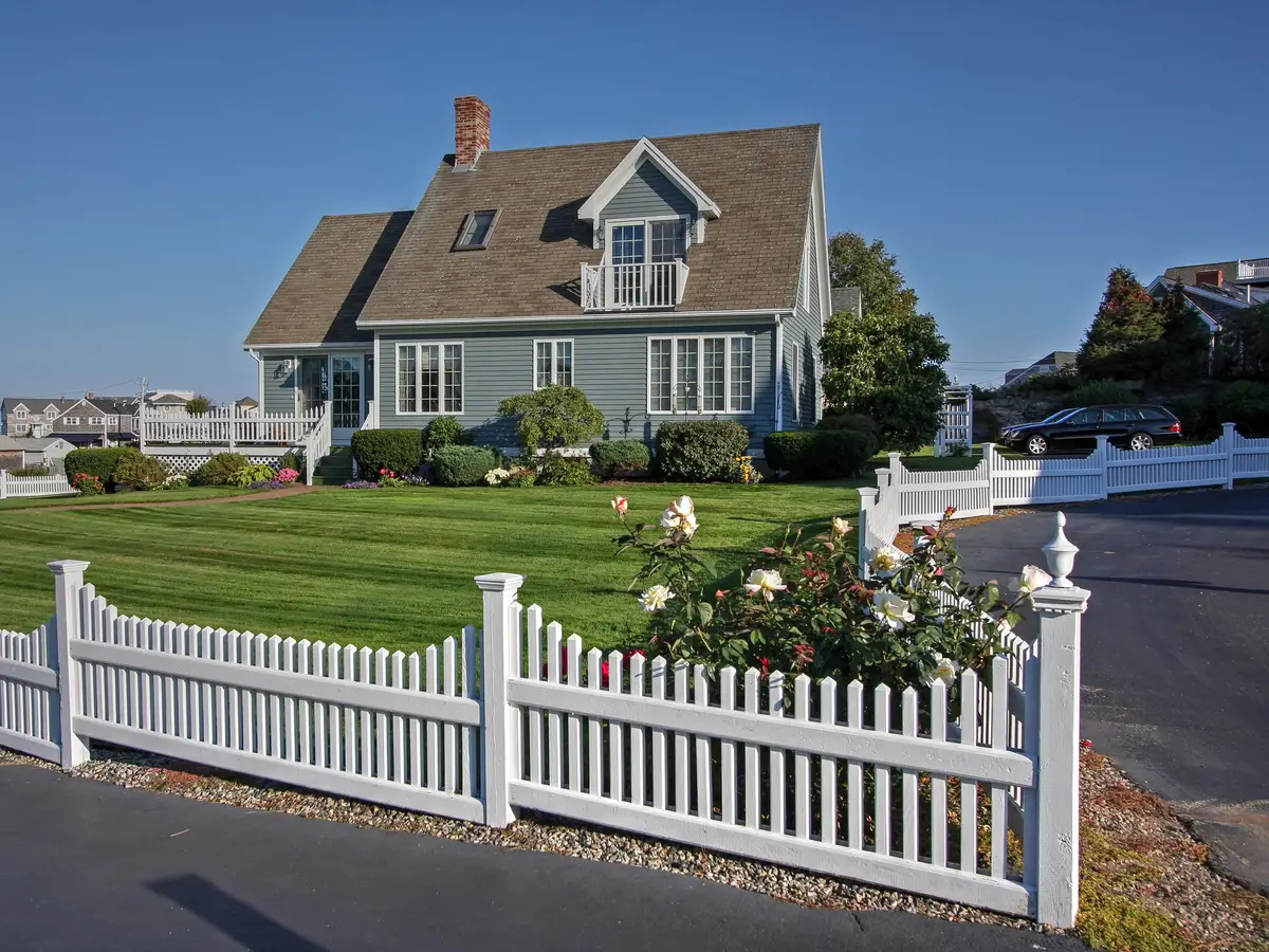 A green lawn behind a white picket fence at a blue-gray house, with rose bushes and a dark car at the right