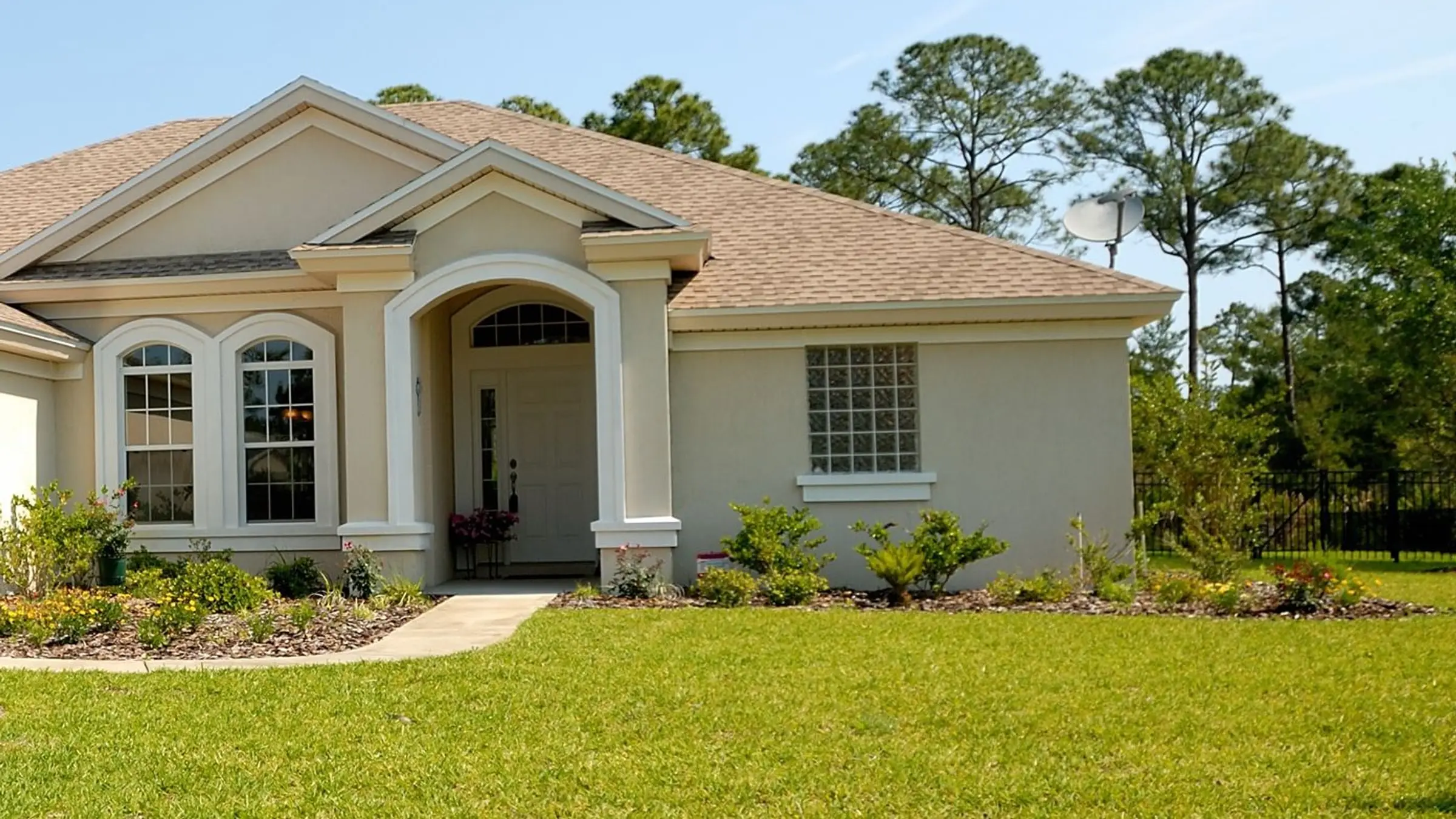 A beige single-story house with a wide green front lawn and pines under a clear blue sky