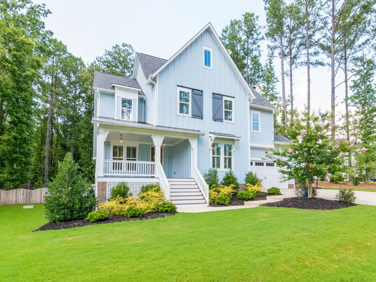 A bright green front lawn in front of a light blue two-story house, with a wooden fence at the left, a driveway, and tall pines
