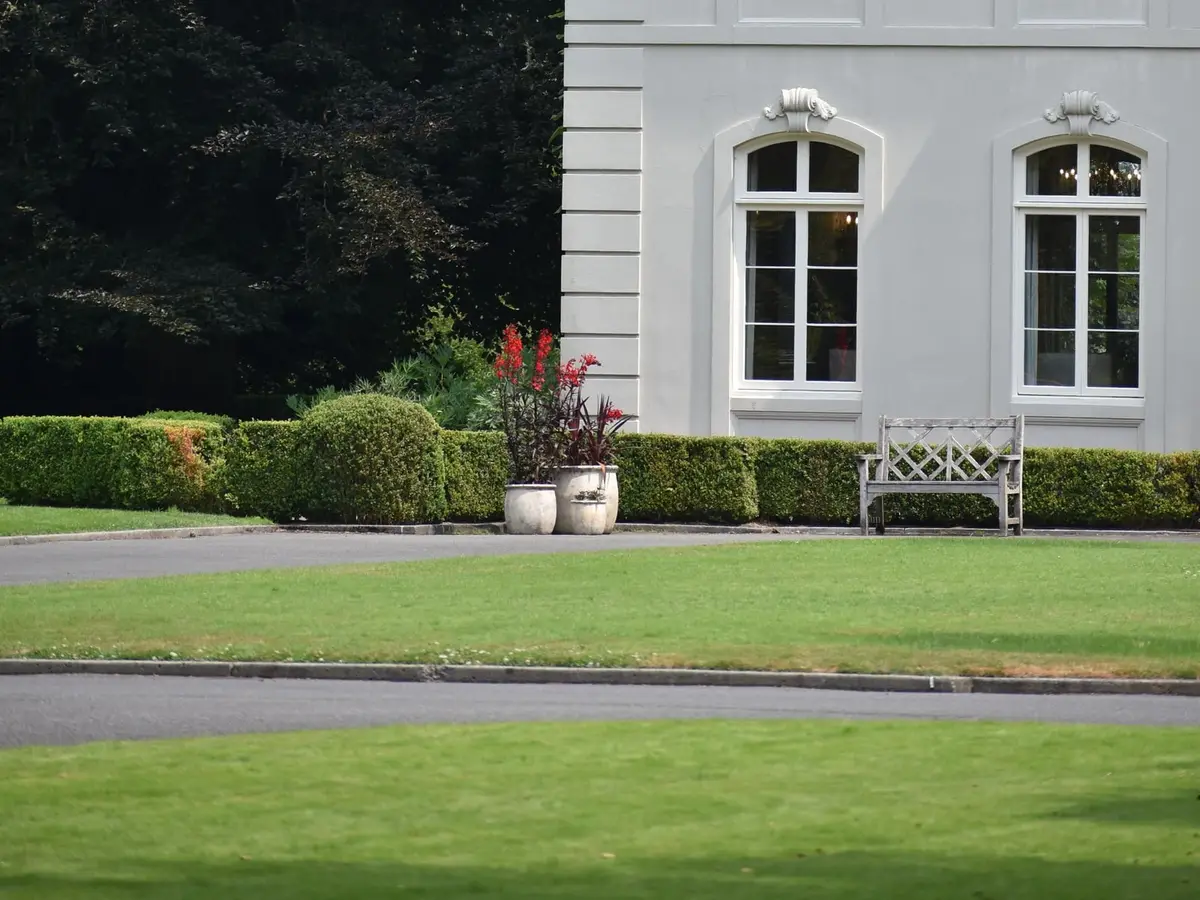 A green lawn and a paved drive in front of a white house, with clipped hedges and a wooden bench