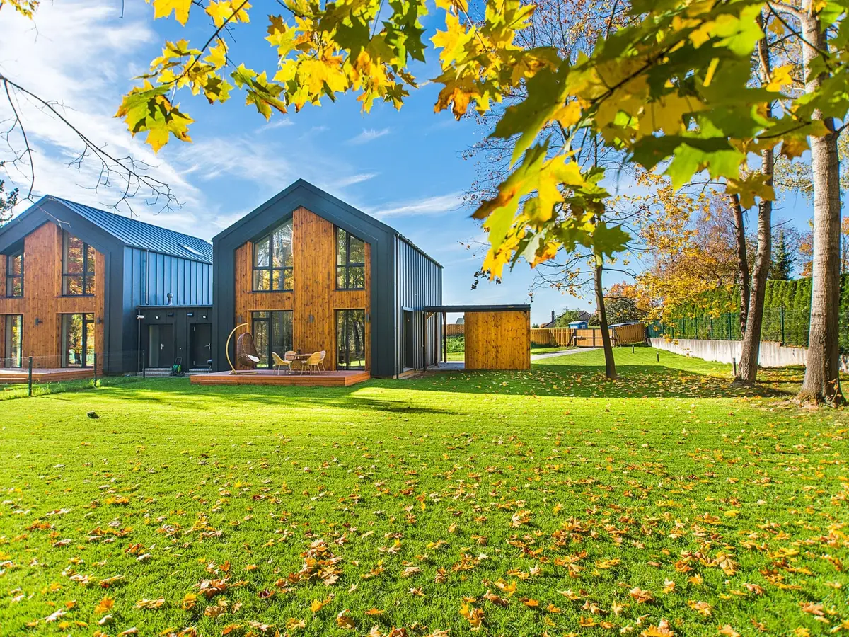 Two wood-and-dark houses beside a green lawn scattered with yellow leaves under a blue sky