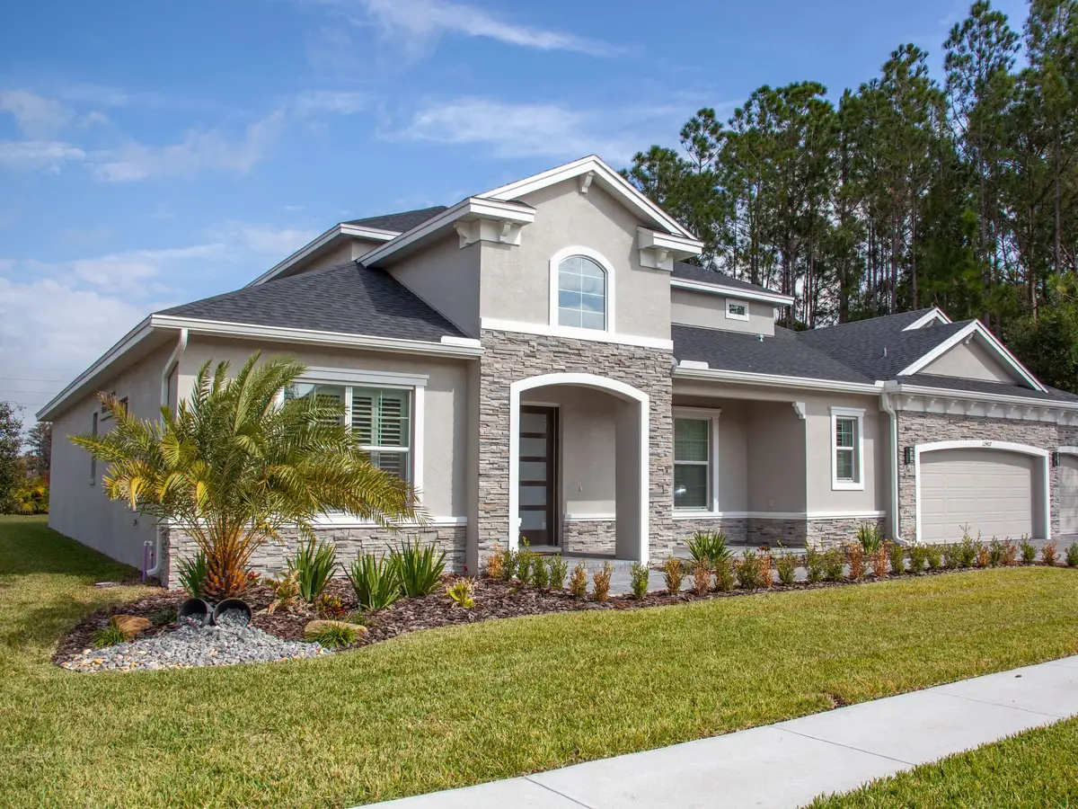 A bright green front lawn, a young palm in a gravel bed, a sidewalk, and a stucco house with a garage and tall pines behind it